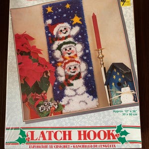 Op de afbeelding: Een boxed latch hook kit met een ontwerp van drie sneeuwmannen met hoeden en sjaals tegen een blauwe achtergrond met gele sterren. De kit bevat de tekst "Latch Hook" en afmetingen van ongeveer 30 x 90 cm.