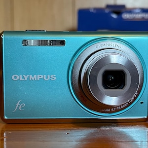May include: A teal Olympus fe digital camera with a silver lens and the Olympus logo. The camera has a zoom lens with a 4.7-18.6mm focal length. The camera is on a wooden surface.