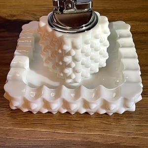 Pode incluir: Um cinzeiro branco com textura de hobnail com um isqueiro correspondente com textura de hobnail. O cinzeiro tem formato retangular com borda ondulada. O isqueiro é prateado.