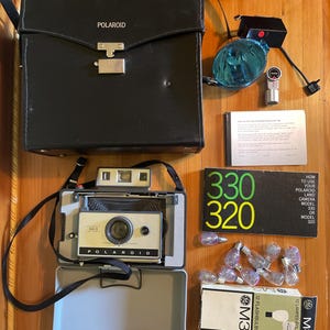 Puede incluir: Cámara Polaroid Land Camera Modelo 320 vintage y accesorios, que incluyen una funda negra, flash y bombillas. La cámara es plateada y negra, con el logotipo de Polaroid. La caja de bombillas dice "M3 12 FLASHBULBS".