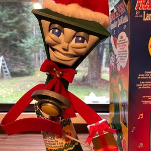 May include: A green and white Christmas lamp post decoration with a red Santa hat and bow. It features a smiling face with blue eyes and holds a small gift box. The text on the decoration says "SING! The Words! I Am Sound Activated."