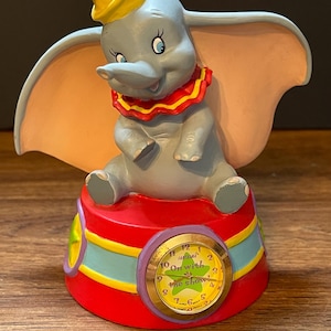 Può includere: Statuina in ceramica di Dumbo l'elefante, con un cappello giallo e un colletto rosso a balze. Dumbo è seduto su una base rossa con un quadrante che dice "On with the show". La statuina è dipinta in grigio, rosa e giallo.