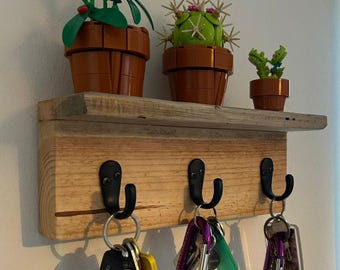 May include: A wooden key rack with four hooks, featuring a rustic design. The rack has a shelf on top, decorated with three small potted plants, including a cactus and a red flower.