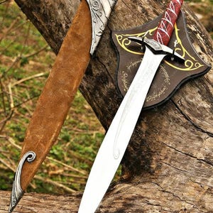 May include: A decorative sword with a silver blade and a red and white handle rests on a wooden surface. The sword is displayed with a brown leather sheath and a brown shield with gold accents. The text "VALYRIAN" is visible.