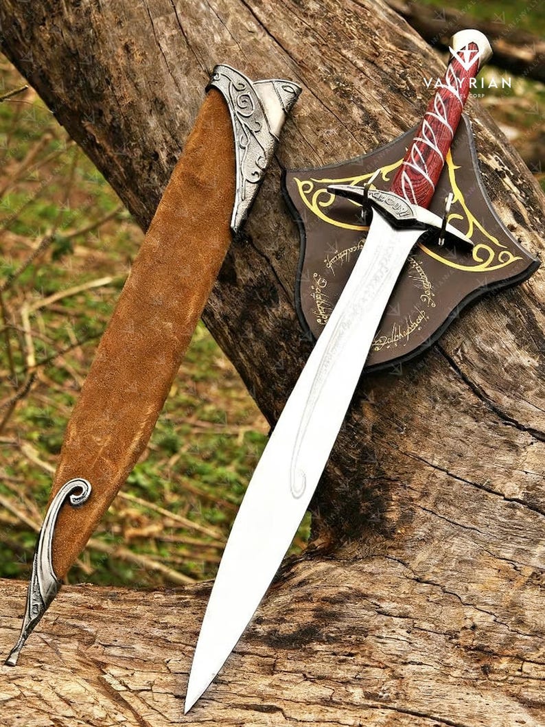 May include: A decorative sword with a silver blade and a red and white handle rests on a wooden surface. The sword is displayed with a brown leather sheath and a brown shield with gold accents. The text "VALYRIAN" is visible.
