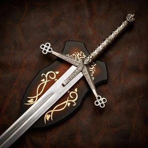 Può includere: Una spada in metallo color argento con un'impugnatura finemente incisa e una guardia a croce con terminali a quattro cerchi. La spada è montata su una placca di legno scuro a forma di scudo, decorata con motivi a foglie dorate intrecciate.