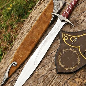 May include: A silver sword with a red and white handle, resting on a wooden surface. The sword has a long, sharp blade and is accompanied by a brown sheath. A brown shield with gold lettering is also present. The text "VALYAN" is visible.
