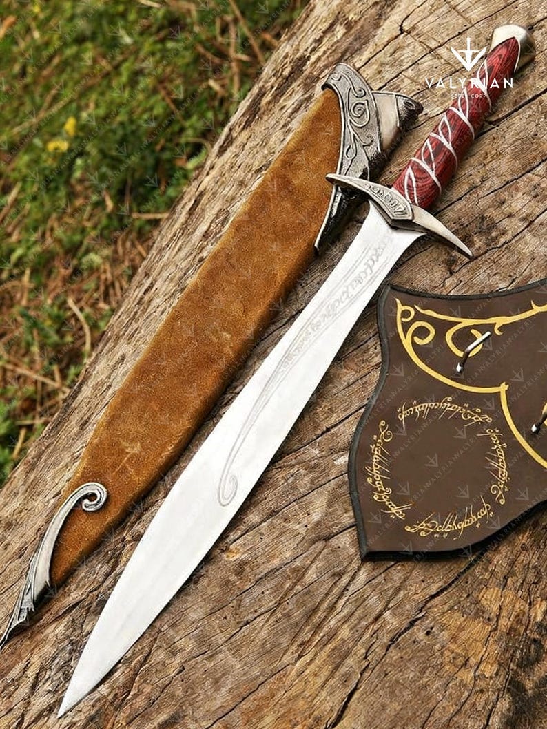 May include: A silver sword with a red and white handle, resting on a wooden surface. The sword has a long, sharp blade and is accompanied by a brown sheath. A brown shield with gold lettering is also present. The text "VALYAN" is visible.