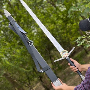 May include: A silver sword with a long, double-edged blade and a black handle is displayed next to its black leather sheath. The sword features a decorative crossguard and a circular emblem with red and green accents.