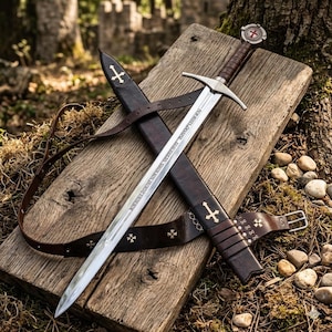 Op de afbeelding: Een zilverkleurig stalen zwaard met een bruin, met leer omwikkeld handvat en een ronde pommel met een rood kruisembleem. Het lemmet is gegraveerd met Latijnse tekst. Het wordt geleverd met een donkerbruine lederen schede met bijpassende riem.