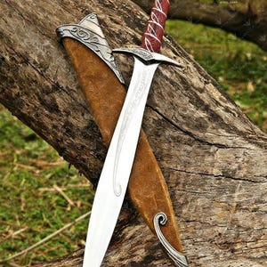 May include: A silver and steel sword with a red and white wrapped handle, resting in a brown leather sheath. The blade has etched designs. The sword is displayed on a weathered wooden log.