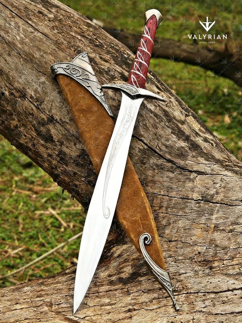 May include: A silver and steel sword with a red and white wrapped handle, resting in a brown leather sheath. The blade has etched designs. The sword is displayed on a weathered wooden log.
