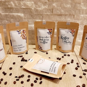Könnte beinhalten: Sechs braune Papiertüten mit verschiedenen Kaffeethemen-Designs und Texten. Die Tüten liegen auf einer Holzoberfläche mit Kaffeebohnen, die um sie herum verstreut sind. Der Text auf den Tüten lautet: "Guter Kaffee, gute Stimmung, guter Tag", "Ruhe bewahren und Kaffee trinken", "Kaffee & Montag" und "So gut, um den Tag zu beginnen".