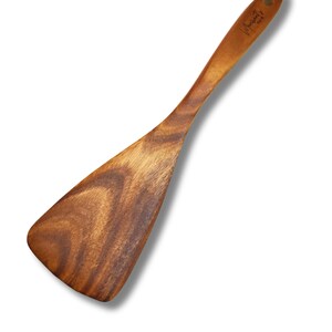 Op de afbeelding: Een houten spatel met een brede, platte kop en een lange steel. De spatel is gemaakt van lichtbruin hout met een natuurlijk nerfpatroon. De steel heeft een klein gaatje om op te hangen en een handtekening.