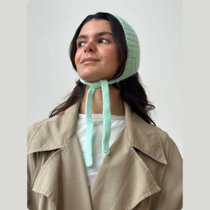 May include: A light green knitted bonnet with long ties. The bonnet is made of a soft, textured yarn and is tied under the chin. The person is wearing a tan trench coat and a white shirt.