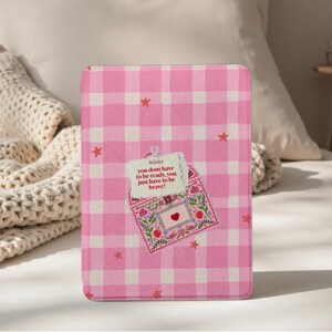 May include: A tablet case with a pink and white gingham pattern, featuring a decorative envelope design. The envelope includes a heart, flowers, and the text "you dont have to be ready, you just have to be brave!" Red star accents are scattered across the case.