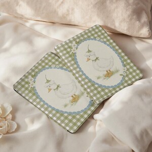 May include: Two rectangular coasters with a green and white gingham pattern border. Each coaster features a white duck illustration wearing a bonnet and a green bow, set within a blue oval frame with floral accents. The background is a soft, off-white color.
