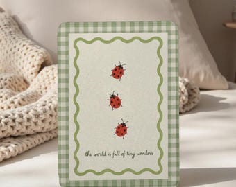 May include: A rectangular card with a green gingham border and a wavy green inner frame. Three red ladybirds with black spots are vertically aligned. The text "the world is full of tiny wonders" is at the bottom.