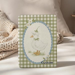 May include: A custom-designed case featuring a charming illustration of a white duck wearing a floral hat and bow. The duck is set within a blue and white oval, surrounded by flowers, against a green and white checkered background. The word "Custom" is at the bottom.