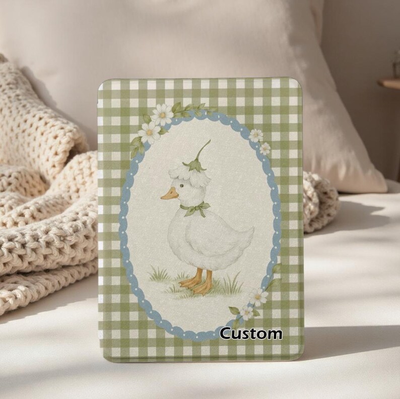May include: A custom-designed case featuring a charming illustration of a white duck wearing a floral hat and bow. The duck is set within a blue and white oval, surrounded by flowers, against a green and white checkered background. The word "Custom" is at the bottom.