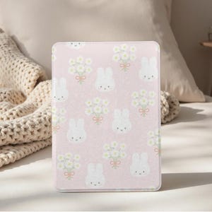 Può includere: Una custodia rosa per tablet con un motivo ripetuto di facce di coniglietti bianchi e bouquet di fiori bianchi legati con nastri rosa. La custodia ha una superficie liscia e una forma rettangolare.