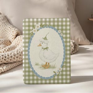 May include: A rectangular case with a green and white gingham pattern. The case features an oval illustration of a white duck wearing a flower hat and a green bow. The duck is standing on grass, surrounded by flowers, with a blue scalloped border.