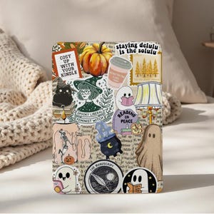 May include: A Kindle case decorated with Halloween-themed stickers. The stickers include a pumpkin, a witch, ghosts, and text that reads "Cosy up with your Kindle" and "Reading in Peace". The case has a white background.