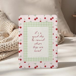 May include: A rectangular card with a pink and white striped border, decorated with red cherry illustrations. The centre has a green gingham pattern, framing the text: "it's a good day to remind others they are loved."