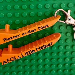 May include: Two orange plastic keychain charms with the text "Retter in der Not" and "AFOLS Little Helper" on a green Lego base.