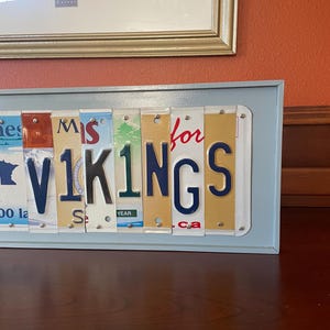 May include: A framed sign made from licence plates spelling out "Miss for Vikings". The sign is in a light blue frame and is sitting on a wooden surface.