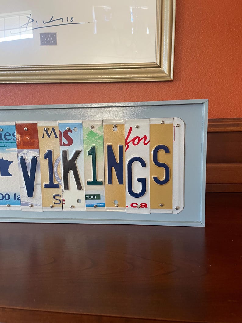May include: A framed sign made from licence plates spelling out "Miss for Vikings". The sign is in a light blue frame and is sitting on a wooden surface.