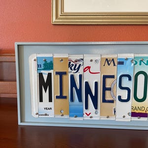 May include: A light blue framed wall art piece featuring the word "MINNESOTA" spelled out using vintage licence plates. The plates are arranged in a row and have various colours and designs. The word "YEAR" is visible on one of the plates.