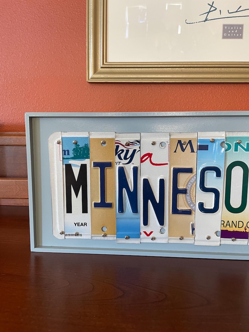 May include: A light blue framed wall art piece featuring the word "MINNESOTA" spelled out using vintage licence plates. The plates are arranged in a row and have various colours and designs. The word "YEAR" is visible on one of the plates.