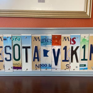 May include: A wall art piece made from vintage number plates spelling out "MINNESOTA VIKINGS". The plates are arranged in a horizontal row and are held together by small metal rivets. The plates are a mix of blue, green, red, and white.