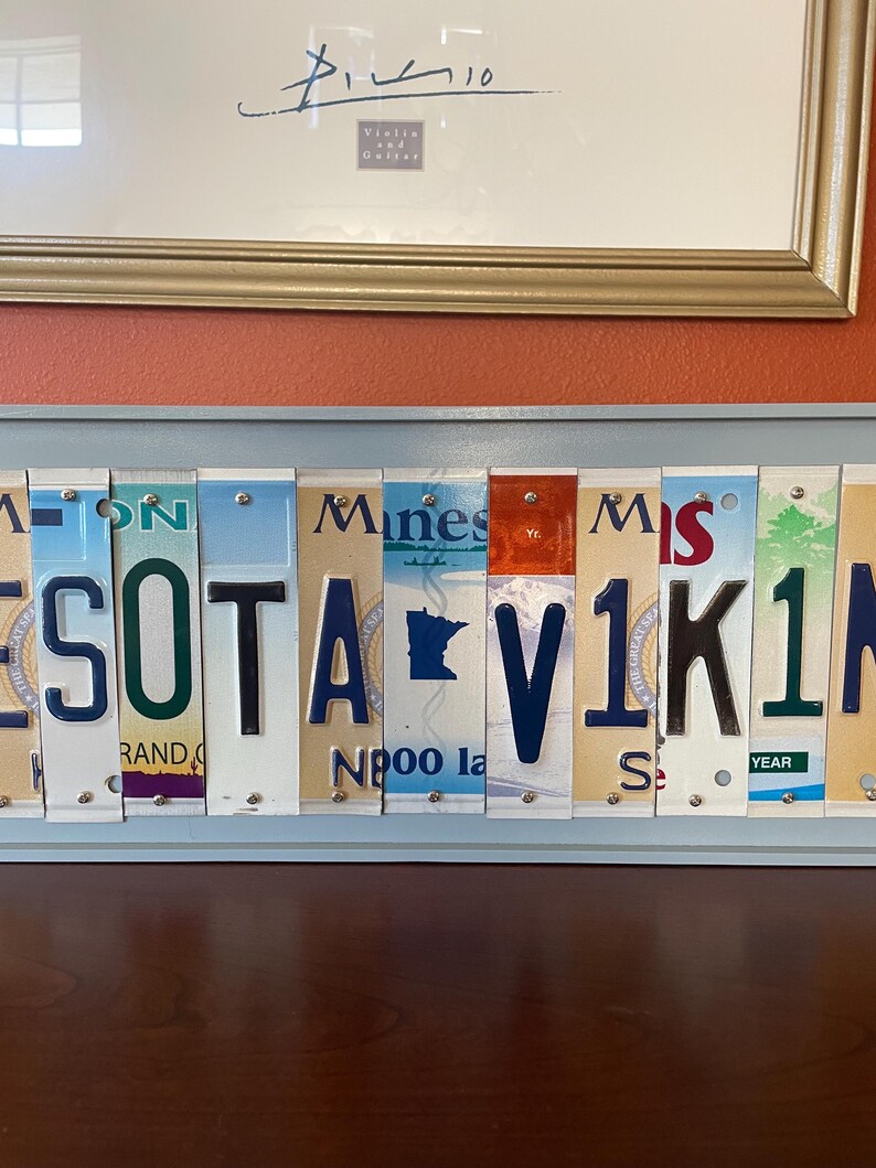 May include: A wall art piece made from vintage number plates spelling out "MINNESOTA VIKINGS". The plates are arranged in a horizontal row and are held together by small metal rivets. The plates are a mix of blue, green, red, and white.