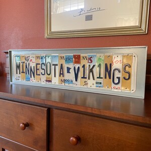 May include: A framed sign made of vintage number plates spelling out "MINNESOTA VIKINGS". The sign is on a wooden surface.