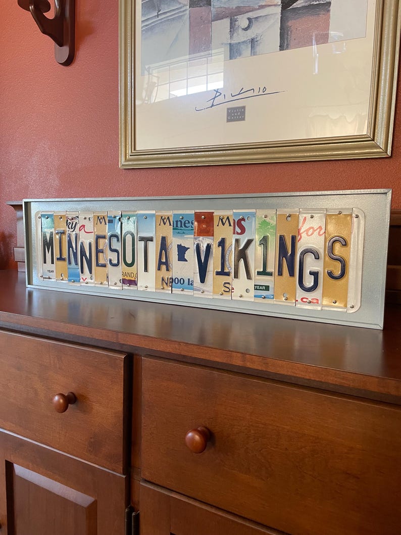 May include: A framed sign made of vintage number plates spelling out "MINNESOTA VIKINGS". The sign is on a wooden surface.