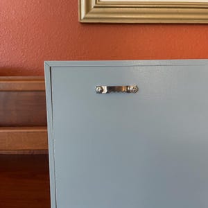 May include: A light blue wooden box with a silver metal handle. The box is closed and has a smooth surface.