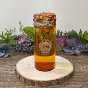 May include: A glass bottle with a layered liquid in shades of orange and yellow, sealed with a brown wax and twine. The bottle has a label with the text "D20". The bottle is displayed on a wooden slice.