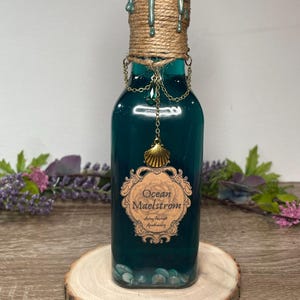 May include: A decorative glass bottle filled with teal liquid, adorned with a rope-wrapped neck and a gold chain with a seashell charm. The bottle features a label that reads "Ocean Maelstrom". The bottle sits on a wooden slice.