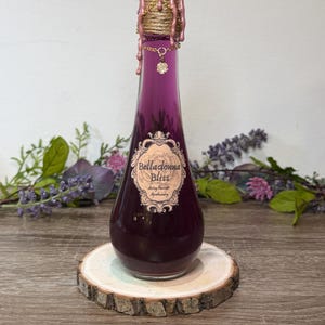 May include: A glass bottle filled with purple liquid featuring a label that reads Belladonna Bliss, Artoy Parrott Apothecary. The neck is wrapped in twine and gold chain, topped with pink wax drips and a small gold charm featuring a flower design.