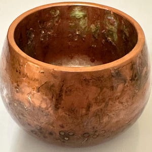 以下が含まれることがあります： 丸みを帯びた形状の手作りの銅色のボウル。ボウルは、明るい銅色から暗い銅色まで、色のバリエーションがあるテクスチャ加工された表面を持っています。内部には緑と茶色のヒントが見られます。ボウルの直径は約10cmです。