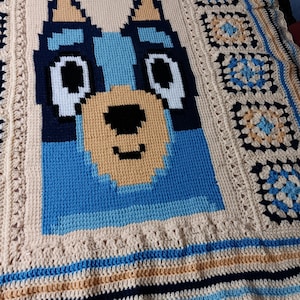 May include: A handmade crochet blanket featuring a cartoon dog face in shades of blue, tan, black, and white. The blanket has a cream-colored background with a border of blue, tan, and navy stripes, and a scalloped edge.