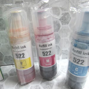 Op de afbeelding: Drie flessen navulinkt, elk in een doorzichtige plastic verpakking. De flessen zijn gelabeld met de tekst "Refill ink" en "Replacement for: 522". De inktkleuren zijn geel, magenta en cyaan.