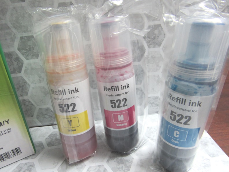 Op de afbeelding: Drie flessen navulinkt, elk in een doorzichtige plastic verpakking. De flessen zijn gelabeld met de tekst "Refill ink" en "Replacement for: 522". De inktkleuren zijn geel, magenta en cyaan.