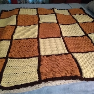 May include: A crocheted blanket with a checkered pattern in shades of brown, cream, and white. The blanket has a brown border and a white lace trim.