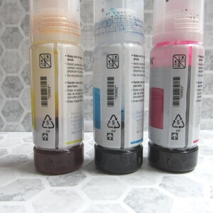 Op de afbeelding: Drie inktflessen met gele, blauwe en roze inkt. Elke fles heeft een zwarte basis en een doorzichtige plastic behuizing met een etiket. De etiketten bevatten tekst en een streepjescode.