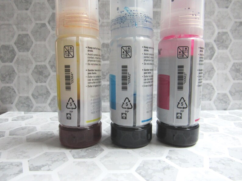 Op de afbeelding: Drie inktflessen met gele, blauwe en roze inkt. Elke fles heeft een zwarte basis en een doorzichtige plastic behuizing met een etiket. De etiketten bevatten tekst en een streepjescode.