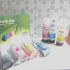 Op de afbeelding: Een groene doos met het opschrift "Refill Ink" en een generieke inktfles, samen met verschillende inktflessen in verschillende kleuren. De flessen zijn voorzien van het opschrift "Epson 522" en bevatten zwarte, magenta, cyaan en gele inkt.