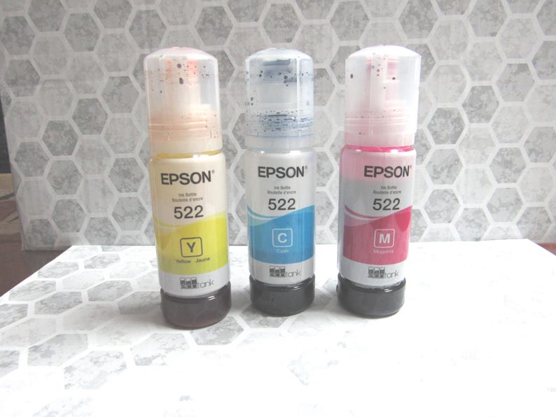 Op de afbeelding: Drie Epson inktflessen, gelabeld met 522, in geel, cyaan en magenta. Elke fles heeft een doorzichtige bovenkant en een zwarte basis. De gele fles is gelabeld met "Y", de cyaan fles is gelabeld met "C", en de magenta fles is gelabeld met "M".